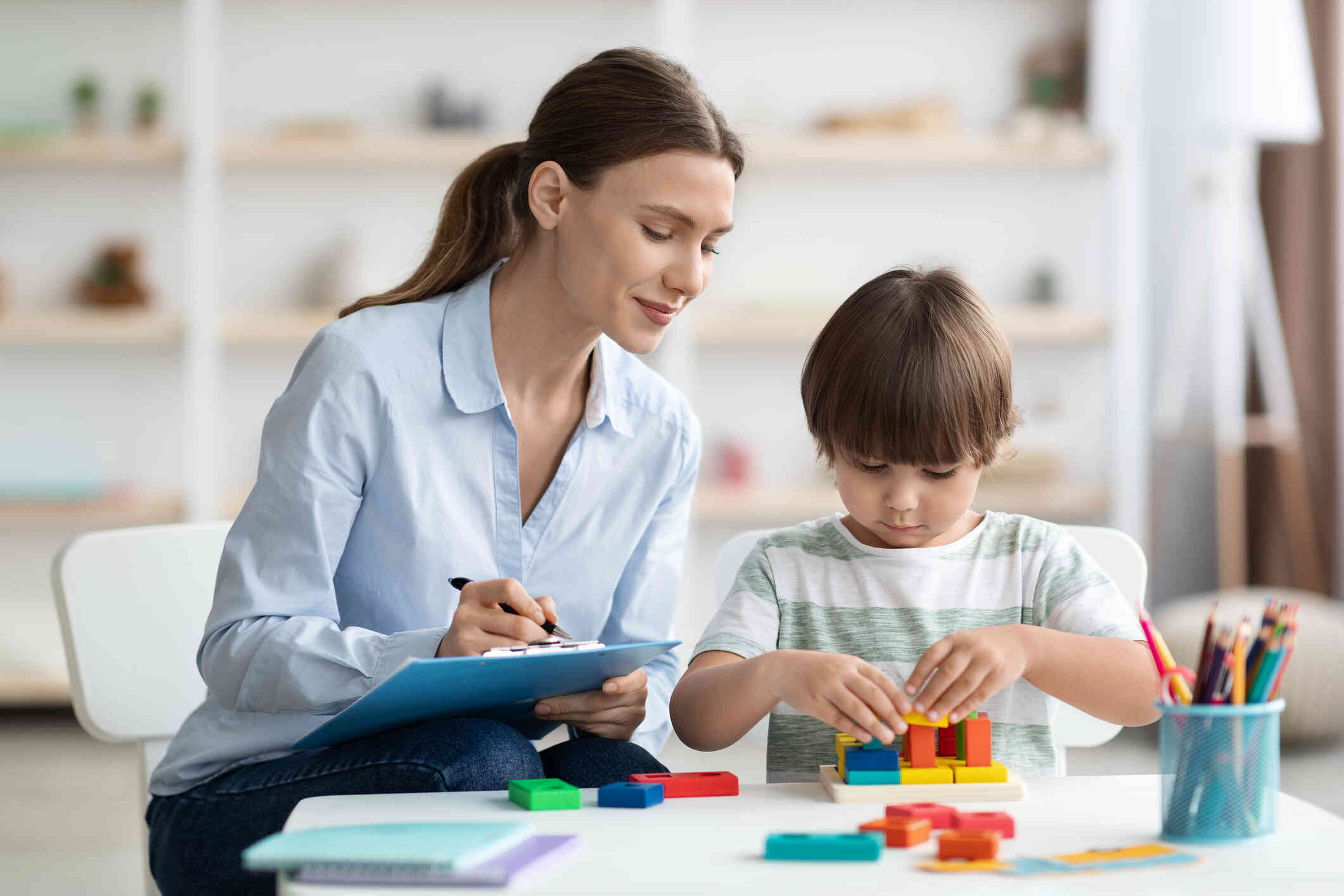 A woman with a clipboard observes a young boy playing with some blocks while smiling softly.